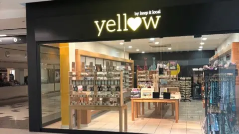 Keep It Local A shop inside a shopping centre that has a black front with a large wall of glass window. there is a yellow sign that reads "yellow, by keep it local" and the "o" of yellow is in the shape of a heart 