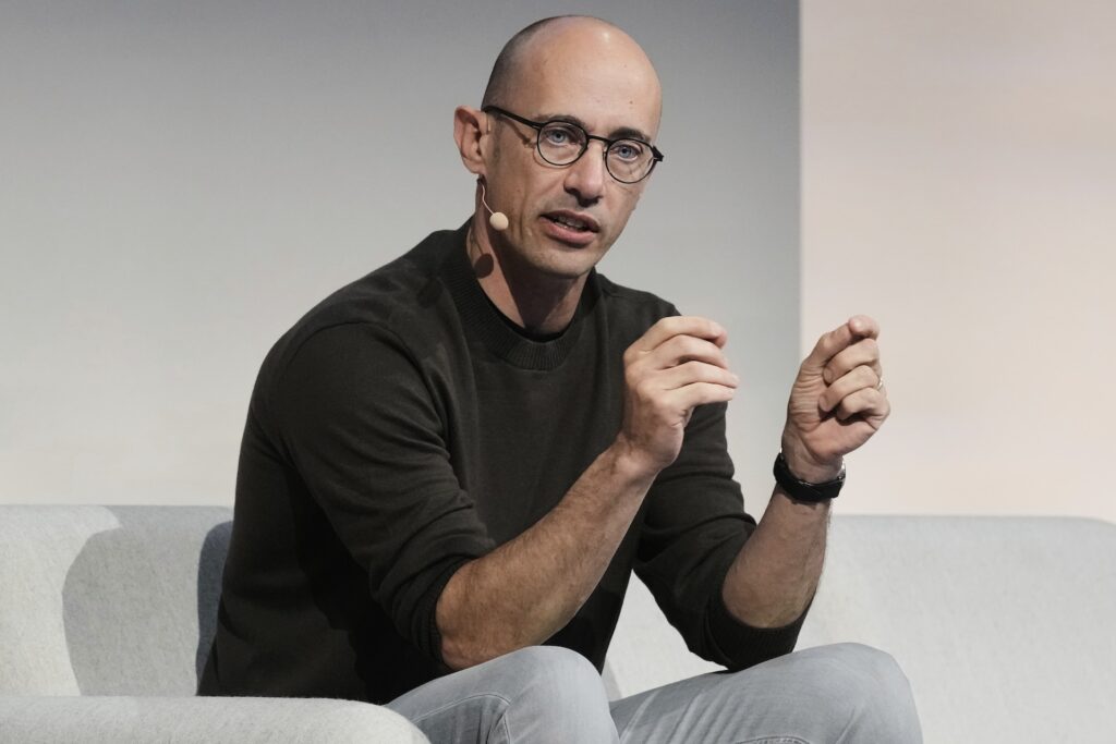 Tobias Lutke in a black shirt and grey jeans sitting on a couch, gesturing with both hands pinching the air as he speaks