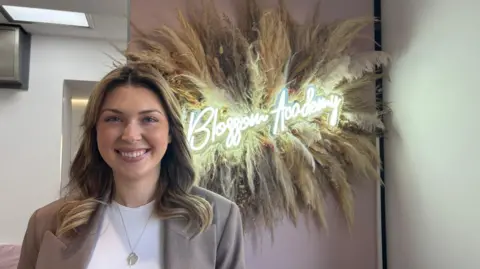 BBC A woman with blonde and brown hair, wearing a white top and light brown blazer. She is standing in front of a pink wall, with a large display of pampas branches. There is a light in the middle that reads "Blossom Academy"