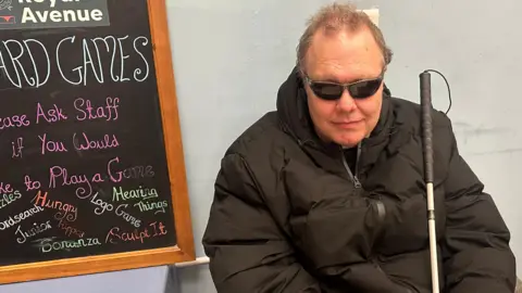 BBC Dave Cunningham is wearing dark glasses and a black coat, and has a white cane in his hand. He is sitting sitting against a grey wall. There is a blackboard on one side and a bookcase on the other.