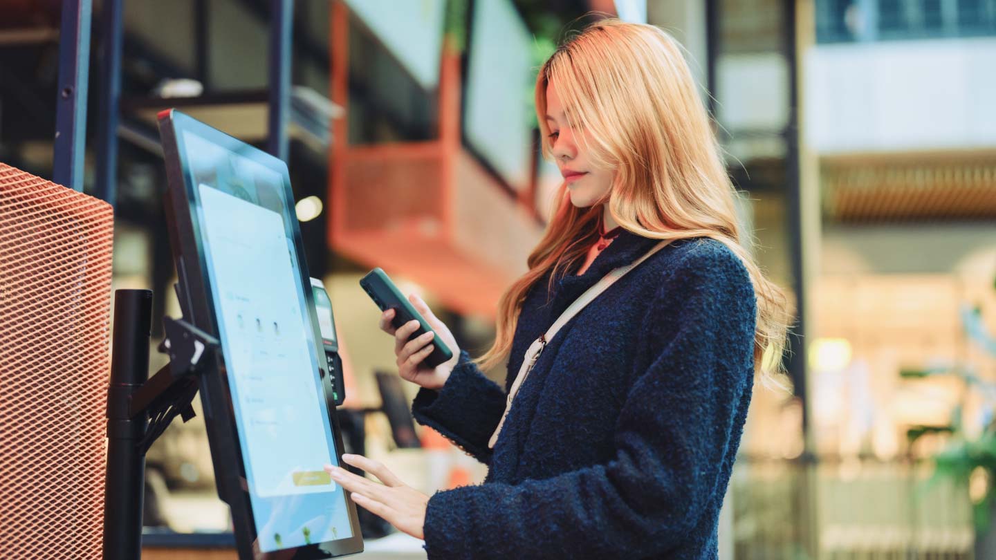 A woman uses a touchscreen kiosk while holding a smartphone in a modern indoor setting.