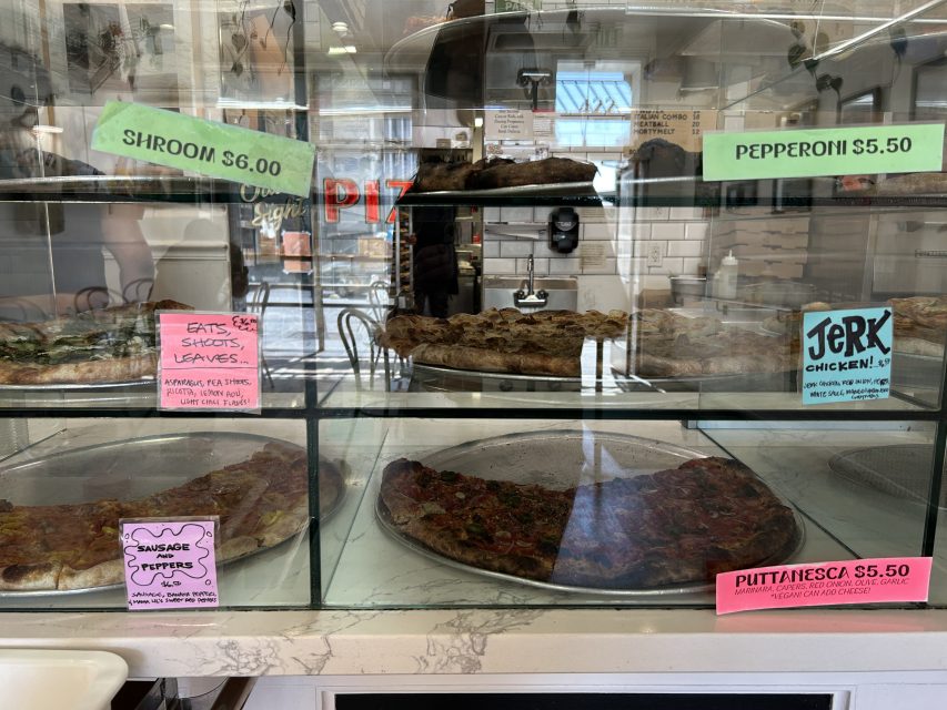 Glass display case with various pizza slices labeled with names and prices, including Shroom, Pepperoni, Sausage Peppers, Jerk Chicken, and Puttanesca.