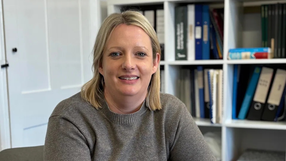 A woman wearing a grey jumper who is smiling at the camera. She has blonde hair and sitting in a room with a bookshelf behind her.