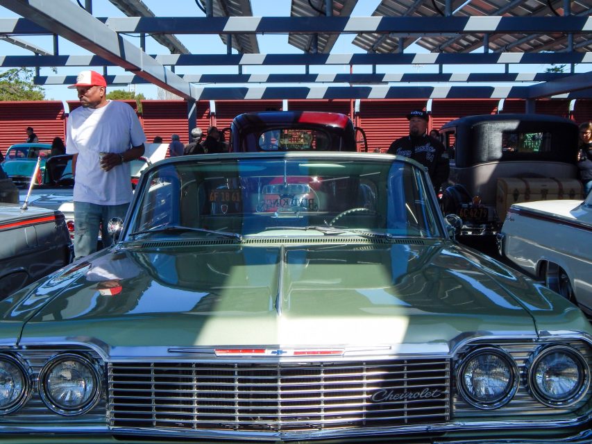 A classic green Chevrolet car is parked at a car show under a partially shaded structure, with people standing and other vintage vehicles in the background.