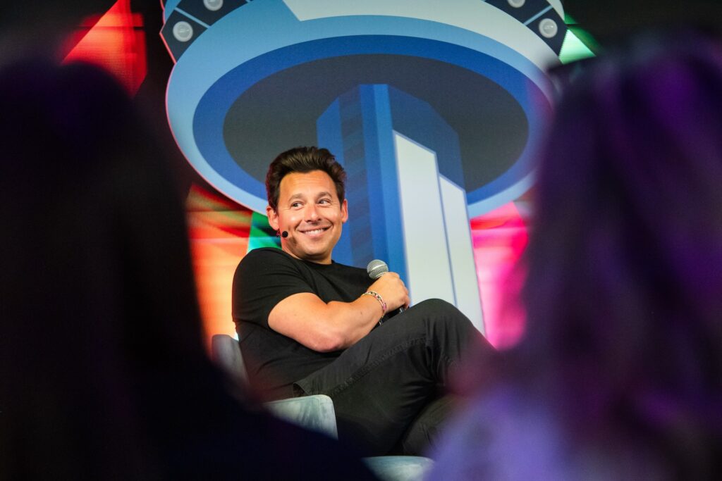 Shopify president Harley Finkelstein smiling as he sits on stage at an event, his arms crossed. He holds a microphone in his hand and wears another microphone on a headset.